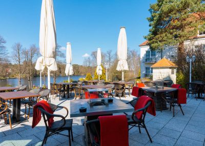 Sonnige Terrasse des Residenz Seehotel mit Blick auf den ruhigen See, ausgestattet mit eleganten Holztischen und komfortablen Stühlen, ideal für ein entspanntes Essen im Freien in naturschöner Umgebung.