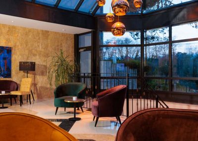 Stimmungsvolle Abendatmosphäre in der Lobby des Residenz Seehotel mit luxuriösen, samtbezogenen Sesseln in tiefen Farbtönen, eleganter Beleuchtung durch moderne Hängelampen und großzügigen Glasfenstern, die einen Blick in den nächtlichen Garten gewähren.